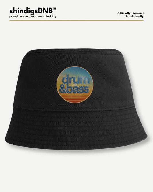 A black bucket hat with a circular retro-style graphic on the front featuring the words 'drum & bass' in blue over a yellow, orange, and blue sunset gradient. The hat is displayed against a light gray background with branding text at the top.